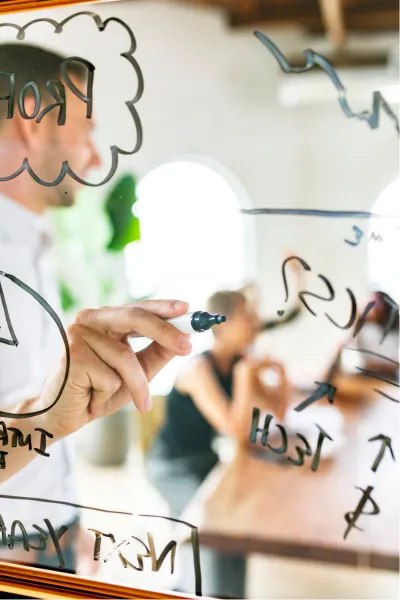 A man writes on a glass wall facilitating a planning session for a group as an executive operational enhancement consultant